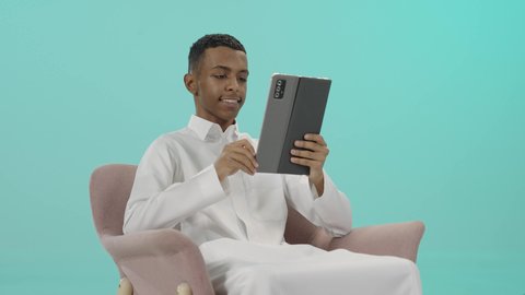 تسلية و استمتاع بالوقت, تصفح الإنترنت وإنجاز المهام عن بعد, بورتريه لرجل عربي خليجي سعودي يرتدي الثوب التقليدي يجلس على كرسي مريح يمسك التابلت بتعابير السعادة, استخدام جهاز تقني حديث تكنولوجي متطور، خلفية زرقاء