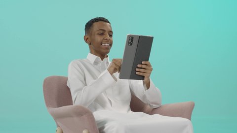 الدردشة مع الأهل والأصدقاء، قضاء وقت ممتع، تصفح الإنترنت وإنجاز المهام عن بعد, بورتريه لرجل عربي خليجي سعودي يرتدي الثوب التقليدي يجلس على كرسي مريح يمسك التابلت بتعابير السعادة, استخدام جهاز تقني حديث تكنولوجي متطور، خلفية زرقاء