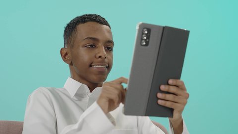 Having fun, surfing the internet and completing tasks remotely, portrait of a Saudi Arabian Gulf man wearing traditional thobe sitting on a comfortable chair holding a tablet with a happy expression, using a modern, advanced technological device, blue background