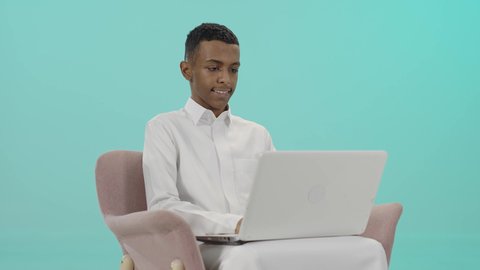 تعابير تدل على الاستمتاع باستخدام الحاسب, دردشة وتواصل عبر الانترنت, استخدام الاجهزة والتقنيات الحديثة والمتطورة, بورتريه لشاب عربي سعودي خليجي مبتسم يرتدي الثوب التقليدي يجلس على كرسي مريح و يمسك بيده اللابتوب, خلفية زرقاء