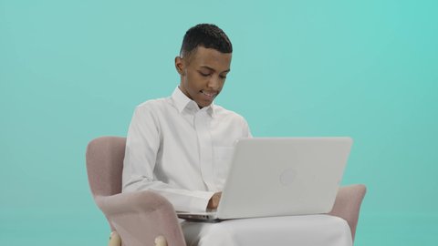 دردشة وتواصل عبر الانترنت, تعابير تدل على الاستمتاع باستخدام الحاسب المحمول, استخدام الاجهزة والتقنيات الحديثة والمتطورة, بورتريه لشاب عربي سعودي خليجي مبتسم يرتدي الثوب التقليدي يجلس على كرسي مريح و يمسك بيده اللابتوب, خلفية زرقاء