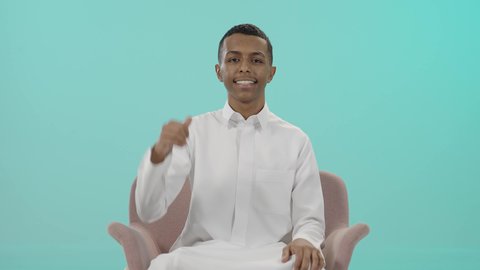 حركة أصبع تدل على القبول والرضا، الإمتياز والتميز والنجاح، بورتريه لرجل عربي خليجي سعودي يرتدي الثوب التقليدي يجلس على كرسي مريح و يرفع أصبع الإبهام لأعلى، إيماءات السعادة والإعجاب والموافقة، خلفية زرقاء