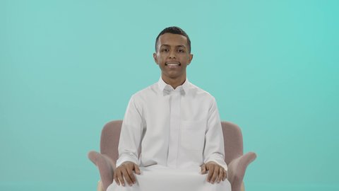 الإمتياز والتميز والنجاح، حركة أصبع تدل على القبول والرضا، بورتريه لرجل عربي خليجي سعودي يرتدي الثوب التقليدي يجلس على كرسي مريح و يرفع أصبع الإبهام لأعلى، إيماءات السعادة والإعجاب والموافقة، خلفية زرقاء