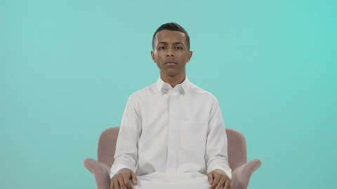 عدم الإعجاب وعدم قبول شيء ما، التقييم السلبي والفشل، بورتريه لرجل عربي خليجي سعودي يرتدي الثوب التقليدي يجلس على كرسي مريح و يرفع إبهامه و يشير للأسفل بانزعاج, عدم الرضا ومنع استخدام شيء معين, خلفية زرقاء