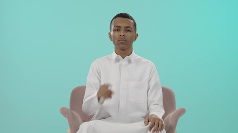 التوقف عن الكلام وخفض الصوت، الهدوء والسكون وعدم الإزعاج، اشارات طلب السكوت والصمت، بورتريه لرجل عربي خليجي سعودي يرتدي الثوب التقليدي و يضع أصبع السبابة على فمه بإيماءات وجه غاضبة، خلفية زرقاء