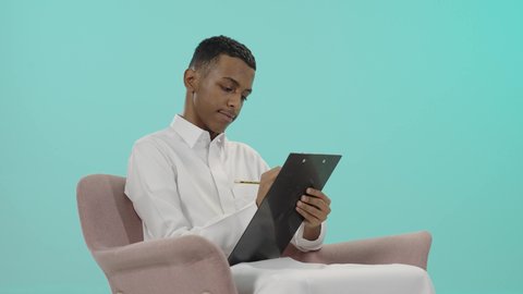 تسجيل المهام والأهداف اليومية، قضاء وقت الفراغ بالتعلم والثقافة، بورتريه لرجل عربي خليجي سعودي يرتدي الثوب التقليدي يجلس على كرسي مريح و يقوم بالكتابة على سنادة ورق سوداء، تعابير التمعن والتركيز خلفية زرقاء