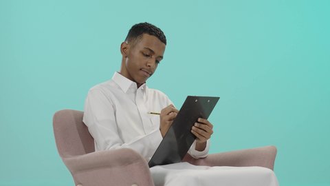 قضاء وقت الفراغ بالتعلم والثقافة، تسجيل المهام والأهداف اليومية، بورتريه لرجل عربي خليجي سعودي يرتدي الثوب التقليدي يجلس على كرسي مريح و يقوم بالكتابة على سنادة ورق سوداء، تعابير التمعن والتركيز خلفية زرقاء