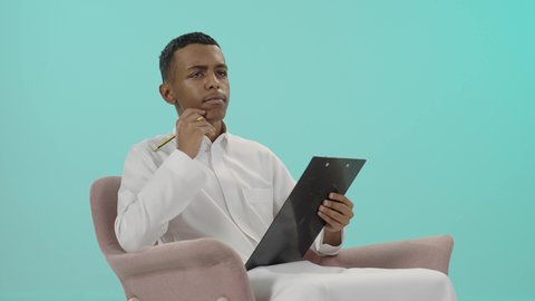 تعابير التفكير لاتخاذ القرارات, أدوات تدوين المهام و الملاحظات, بورتريه لرجل عربي خليجي سعودي يرتدي الثوب التقليدي يجلس على كرسي مريح و يقوم بالكتابة على سنادة ورق سوداء، تعابير التمعن والتركيز، خلفية زرقاء