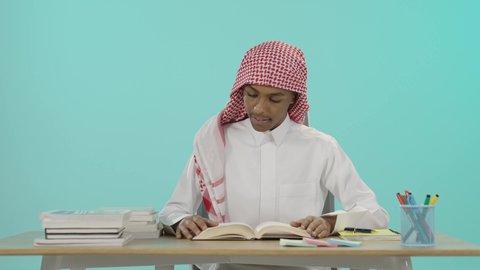The concept of the love of reading and learning, expressions of diligence, seriousness and activity, preparing for the new school year, preparing lessons to achieve success, a portrait of a young Saudi Arabian Gulf man wearing traditional thobe and shemagh sitting at a table reading a book, developing educational curricula in Saudi Arabia, blue background