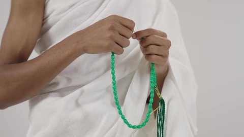 التهليل والتسبيح وذكر الله تعالى، طلب المغفرة والدعاء والصفح، الاجتهاد في العبادة, قراءة الأذكار والأدعية، بورتريه مقرب لحاج عربي خليجي سعودي يرتدي لباس الإحرام يمسك بيده سبحة خضراء، التقرب لرب العالمين، خلفية رمادي