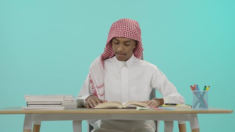 Reading books and gaining knowledge, preparing and reviewing scientific research, the concept of the love of reading and learning, expressions of diligence, seriousness and activity, preparing for the new school year, preparing lessons to achieve success, portrait of a young Saudi Arabian Gulf man wearing traditional thobe and shemagh sitting at a table reading a book, blue background