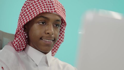 متابعة الدرجات من خلال الحاسب المحمول، تطوير المناهج التعليمية في السعودية، الاجتهاد وتحضير الدروس, بورتريه لشاب عربي خليجي سعودي يرتدي الثوب التقليدي و شماغ يجلس على الطاولة يقوم باستخدام اللابتوب في المذاكرة, خلفية زرقاء