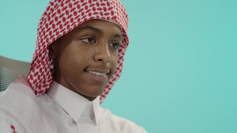 Expressions and gestures of concentration and integration, portrait of a smiling young Saudi Arabian Gulf man wearing traditional thobe and shemagh, immersed in his work, concept of diligence and excellence, preparation for the new school year, blue background