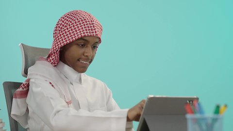 مراجعة الدروس اليومية عبر الانترنت، تطور العلم والتعليم في السعودية,  بورتريه لشاب عربي خليجي سعودي مبتسم يرتدي الثوب التقليدي و الشماغ يقوم باستخدام الجهاز اللوحي, مفهوم الاجتهاد والتفوق, الاستعداد للعام الدراسي الجديد، خلفية زرقاء