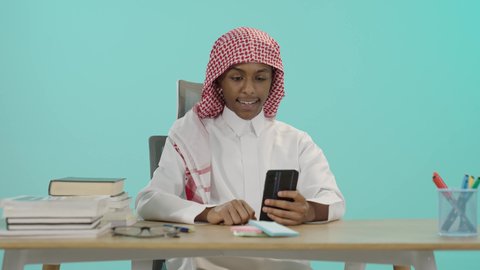 استعراض المنشورات والتحديثات على الإنترنت، البحث والمطالعة من خلال الجوال، تصفح التطبيقات ووسائل التواصل الاجتماعي، استخدام الأجهزة والتقنيات الحديثة والمتطورة, بورتريه لشاب عربي خليجي سعودي مبتسم يرتدي الثوب التقليدي و الشماغ يجلس على الطاولة يمسك بيده الهاتف المحمول، خلفية زرقاء