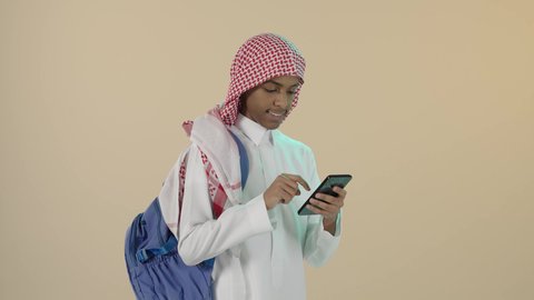البحث والمطالعة من خلال الجوال، تصفح التطبيقات ووسائل التواصل الاجتماعي، استخدام الأجهزة والتقنيات الحديثة والمتطورة, بورتريه لشاب عربي خليجي سعودي مبتسم يرتدي الثوب التقليدي و الشماغ و حقيبة ظهر يستخدم الهاتف المحمول, خلفية بيج