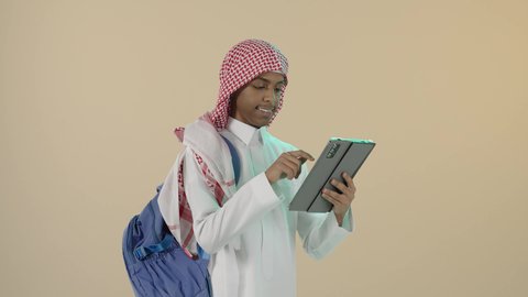 استخدام التقنيات الحديثة في التعليم والتدريس عن بعد، موسم العودة للدراسة, قضاء وقت ممتع على الإنترنت، اللعب على الجهاز اللوحي، بورتريه لشاب عربي خليجي سعودي مبتسم يرتدي الثوب التقليدي و الشماغ و حقيبة ظهر يقوم بتصفح التابلت, خلفية بيج