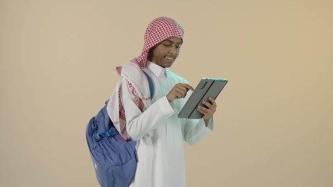 طرائق تعليمية جديدة متطورة، استخدام الأجهزة التقنية الحديثة والمتطورة في المجال الدراسي، قضاء وقت ممتع على الإنترنت، اللعب على الجهاز اللوحي، بورتريه لشاب عربي خليجي سعودي مبتسم يرتدي الثوب التقليدي و الشماغ و حقيبة ظهر يقوم بتصفح التابلت, خلفية بيج