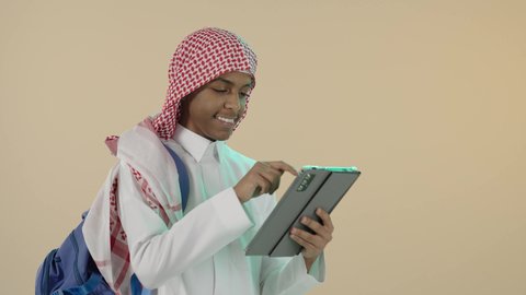 Using modern and advanced technical devices in the academic field, spending fun time on the Internet, playing on the tablet, portrait of a smiling Saudi Arabian Gulf young man wearing traditional thobe, shemagh and backpack browsing the tablet, beige background