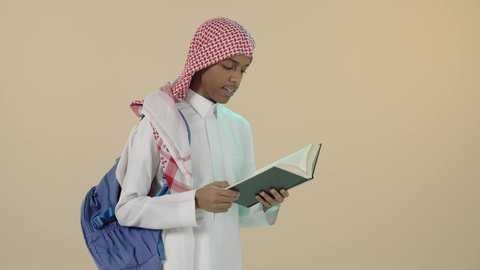 Preparing for the new school year, reviewing daily lessons, preparing lessons to achieve success, portrait of a young Saudi Arabian Gulf man wearing traditional thobe, shemagh and backpack reading a book, developing educational curricula in Saudi Arabia, beige background