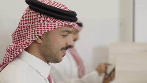 The use of modern technology, a close-up shot of a young Arab Gulf Saudi man wearing traditional attire and a ghutrah, looking at something with gestures of engagement and focus, a man browsing a mobile phone, accomplishing work tasks online.