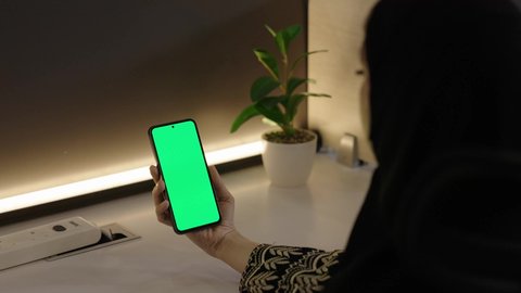 The use of modern technology to accomplish tasks remotely, watching favorite programs, displaying a blank green screen on technical devices, a close-up shot from behind of a Saudi Gulf Arab woman holding a mobile phone.