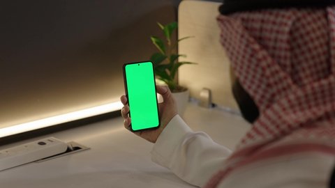 عرض شاشة خضراء فارغة على الأجهزة التقنية، استخدام التكنولوجيا الحديثة في انجاز المهام عن بعد، مشاهدة البرامج المفضلة، تصوير مقرب من الخلف لرجل عربي خليجي سعودي يرتدي الزي التقليدي و الشماغ الهاتف المحمول