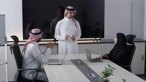 A group of Arab Gulf Saudi employees wearing traditional attire are engaged in discussions to achieve strategies and goals inside the meeting room, collaborating with colleagues to accomplish work tasks, exchanging experiences and skills, enjoying a comfortable work environment in Saudi companies, using modern technological devices, and developing and studying new projects.