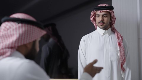 Two Arab Gulf men wearing traditional attire and a ghutrah are sitting inside an office discussing work, developing the business and solving problems, distributing work tasks, developing company goals, in a Saudi work environment.
