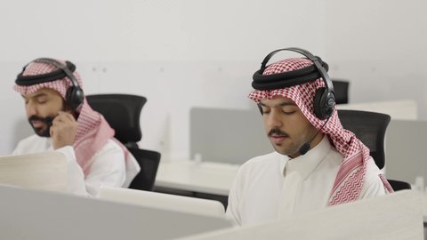 العمل في المكتب عن طريق الحاسوب المحمول, متابعة وتطوير العمل في الشركات السعودية, موظفون عربيون خليجيون سعودوين يعملون  في خدمة العملاء و يرتدون الزي التقليدي و الشماغ و سماعة الهيدفون للتواصل مع العملاء , الاستفسارات و الرد على الأسئلة, مكالمات فيديو خلال اجتماع العمل