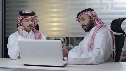 اجتماع ومناقشة عمل في مقر الشركة، رجل أعمال عربي خليجي سعودي يرتدي الثوب التقليدي و الشماغ يقوم بالعمل على الحاسب المحمول مع زميله داخل المكتب، تطوير ودراسة المشاريع الجديدة في شركات المملكة العربية السعودية، استخدام جهاز تقني حديث، جمع البيانات وحل المشكلات
