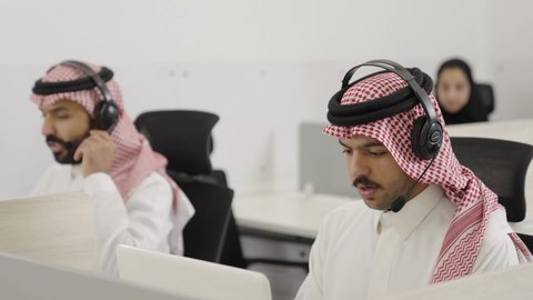 الرد على الاستعلامات والاستفسارات، استخدام التقنيات الحديثة والمتطورة، مكالمات فيديو خلال اجتماع العمل, رجلان عربيان خليجيان سعوديان يرتديان الثوب التقليدي و الشماغ يضعان سماعات الهيدفون يجلسان داخل المكتب يعمل  على الحاسب المحمول فى مجال خدمة العملاء