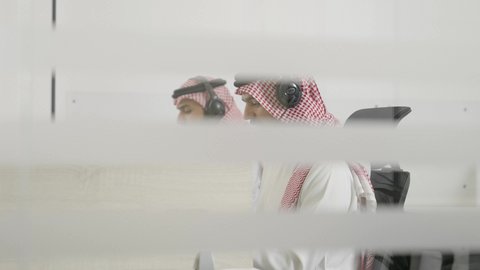 The use of modern and advanced technologies, video calls during a business meeting, two Arab Gulf men from Saudi Arabia wearing traditional thobes and ghutras, wearing headphones, sitting inside the office working on a laptop in the field of customer service, responding to inquiries and questions.