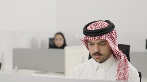 تبادل الخبرات والمهارات، الإشارة بإصبع السبابة إلى شيء ما, مناقشة الخطط والمشاريع, التعاون في إنجاز مهام العمل, استخدام الأجهزة التقنية الحديثة والمتطورة, شاب عربي خليجي سعودي  يرتدي الثوب التقليدي و الشماغ يجلس على المكتب داخل الشركة يقوم باستخدام الحاسب المحمول برفقة زميله