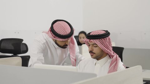Pointing with the index finger at something, exchanging experiences and skills, discussing plans and projects, collaborating to accomplish work tasks, using modern and advanced technical devices, a young Saudi Arabian Gulf man wearing traditional attire and a ghutrah sitting at a desk inside the company using a laptop with his colleague.
