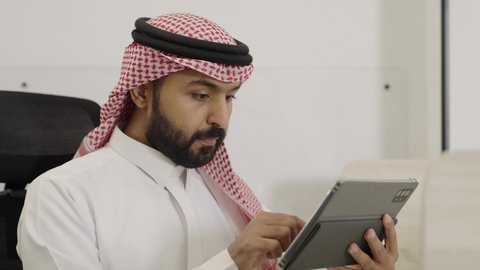 مفهوم إنجاز المشاريع والمهام العمل اونلاين، النقر على شاشة التابلت، استخدام الأجهزة التقنية الحديثة، تصفح مواقع التواصل الاجتماعي، التواصل مع الأهل والأصدقاء عبر الايباد، تصوير مقرب لرجل عربيى خليجي سعودي يرتدي الثوب التقليدي و الشماغ يقوم باستخدام الجهاز اللوحي يجلس فى المكتب بجانب زميلة