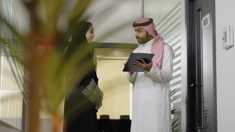 Verification and auditing of financial offers, developing a plan and work mechanism, writing and recording data and notes, completing office tasks, understanding and cooperating among colleagues to achieve goals, an Arab Gulf Saudi man wearing traditional attire and a ghutrah explaining tasks to his female colleague inside the company.