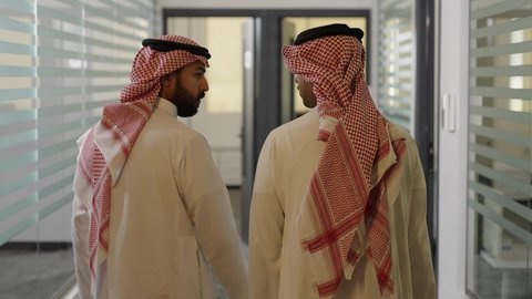 Preparation and readiness to attend the meeting, an administrative office job in the Kingdom of Saudi Arabia, exchanging expertise among the team to achieve the goal, a shot from behind of two Arab Gulf Saudi men wearing traditional thob and ghutrah walking inside the company headquarters.