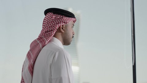 المباني والأبراج التجارية المشهورة، الوقوف باستقامه بتعابير السعادة و السرور، النمو والتطور العمراني، تصوير من الجانب لرجل عربي خليجي سعودي يرتدي الشماغ ينظر من نافذة المكتب على برج المجدول في مدينة الرياض ، الفنون الهندسية المعمارية لناطحات السحاب في المملكة العربية السعودية، معالم الرياض السياحية
