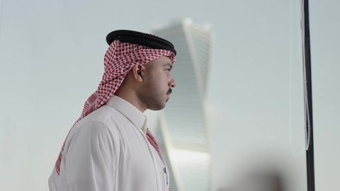 الوقوف باستقامه بتعابير السعادة و السرور، المباني والأبراج التجارية المشهورة، النمو والتطور العمراني، تصوير من الجانب لرجل عربي خليجي سعودي يرتدي الشماغ ينظر من نافذة المكتب على برج المجدول في مدينة الرياض ، الفنون الهندسية المعمارية لناطحات السحاب في المملكة العربية السعودية، معالم الرياض السياحية