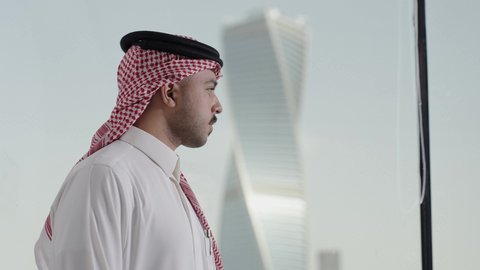 الوقوف بايدي متقاطعه بتعابير الثقة بالنفس، المباني والأبراج التجارية المشهورة، النمو والتطور العمراني، تصوير من الجانب لرجل عربي خليجي سعودي يرتدي الشماغ ينظر من نافذة المكتب على برج المجدول في مدينة الرياض ، الفنون الهندسية المعمارية لناطحات السحاب في المملكة العربية السعودية، معالم الرياض السياحية