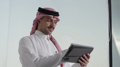 إدارة شؤون الأعمال وتنظيمها عبر الاجهزة التقنية،  دمج العمل مع التقنية والتكنولوجيا، تصفح مواقع التواصل الاجتماعي، تصوير مقرب لرجل اعمال عربي خليجي سعودي يرتدي الزي التقليدي والغترة يعمل على جهاز التابلت المحمول بإيماءات التركيز داخل المكتب