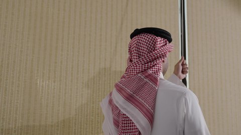 النهضة العمرانية في المدن الخليجية، تصوير من الخلف لرجل عربي خليجي سعودي يرتدي الثوب والشماغ يقف أمام نافذة المكتب يفتح الستائر مطلًا على الأبراج وناطحات السحاب في مدينة الرياض