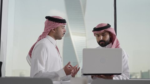 بيئة العمل المريحة في السعودية، تدريب الموظفين الأساليب الجديدة في العمل لتحقيق أهداف الشركة، دمج العمل مع التقنية والتكنولوجيا، زميلان عربيان خليجيان سعوديان يرتديان الزي التقليدي و الشماغ يقومان بإنجاز المهام عبر الحاسوب المحمول، الاشارة الى اللابتوب، العمل الجماعي وروح الفريق الواحد