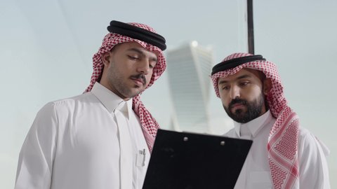 التفاهم والتعاون بين الزملاء ووضع الخطط الاستراتيجية لتحقيق الأهداف، العمل الجماعي وروح الفريق الواحد، ضمان فهم واضح وشامل للاعمال،  تصوير مقرب لموظفان عربيان خليجيان سعوديان يرتديان الثوب التقليدي و الغتره يعقدون اجتماع داخل مقر الشركة، تعلم اساليب العمل والتطوير المهن، تدووين الملاحظات و تسجيلها