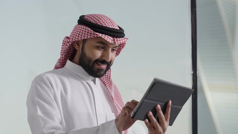 دمج العمل مع التقنية والتكنولوجيا، تصفح مواقع التواصل الاجتماعي، إدارة شؤون الأعمال وتنظيمها عبر الاجهزة التقنية، تصوير مقرب لرجل اعمال عربي خليجي سعودي يرتدي الزي التقليدي والغترة يعمل على جهاز التابلت المحمول بإيماءات التركيز داخل المكتب