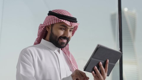 تصفح مواقع التواصل الاجتماعي، إدارة شؤون الأعمال وتنظيمها عبر الاجهزة التقنية، دمج العمل مع التقنية والتكنولوجيا، تصوير مقرب لرجل اعمال عربي خليجي سعودي يرتدي الزي التقليدي والغترة يعمل على جهاز التابلت المحمول بإيماءات التركيز داخل المكتب
