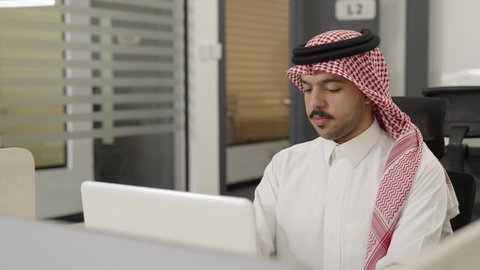 إدارة شؤون الأعمال وتنظيمها عبر الاجهزة التقنية، مهن ووظائف ادارية مكتبية، دمج العمل مع التقنية والتكنولوجيا، رجل عربي خليجي سعودي يرتدي الثوب والشماغ, العمل على الحاسوب المحمول داخل المكتب