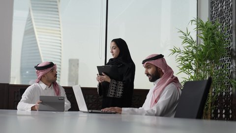 A meeting and work discussion at the company headquarters, using modern technology, developing and studying new projects in the Kingdom of Saudi Arabia, collecting data and solving problems, an Arab Gulf businessman from Saudi Arabia wearing a white thobe and a ghutrah working on a laptop with his colleagues inside the office.