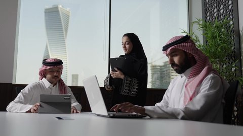 استخدام جهاز تقني حديث، تطوير ودراسة المشاريع الجديدة في المملكة العربية السعودية، جمع البيانات وحل المشكلات، اجتماع ومناقشة عمل في مقر الشركة، رجل أعمال عربي خليجي سعودي يرتدي الثوب الابيض و الشماغ يقوم بالعمل على الحاسب المحمول مع زملاءه داخل المكتب